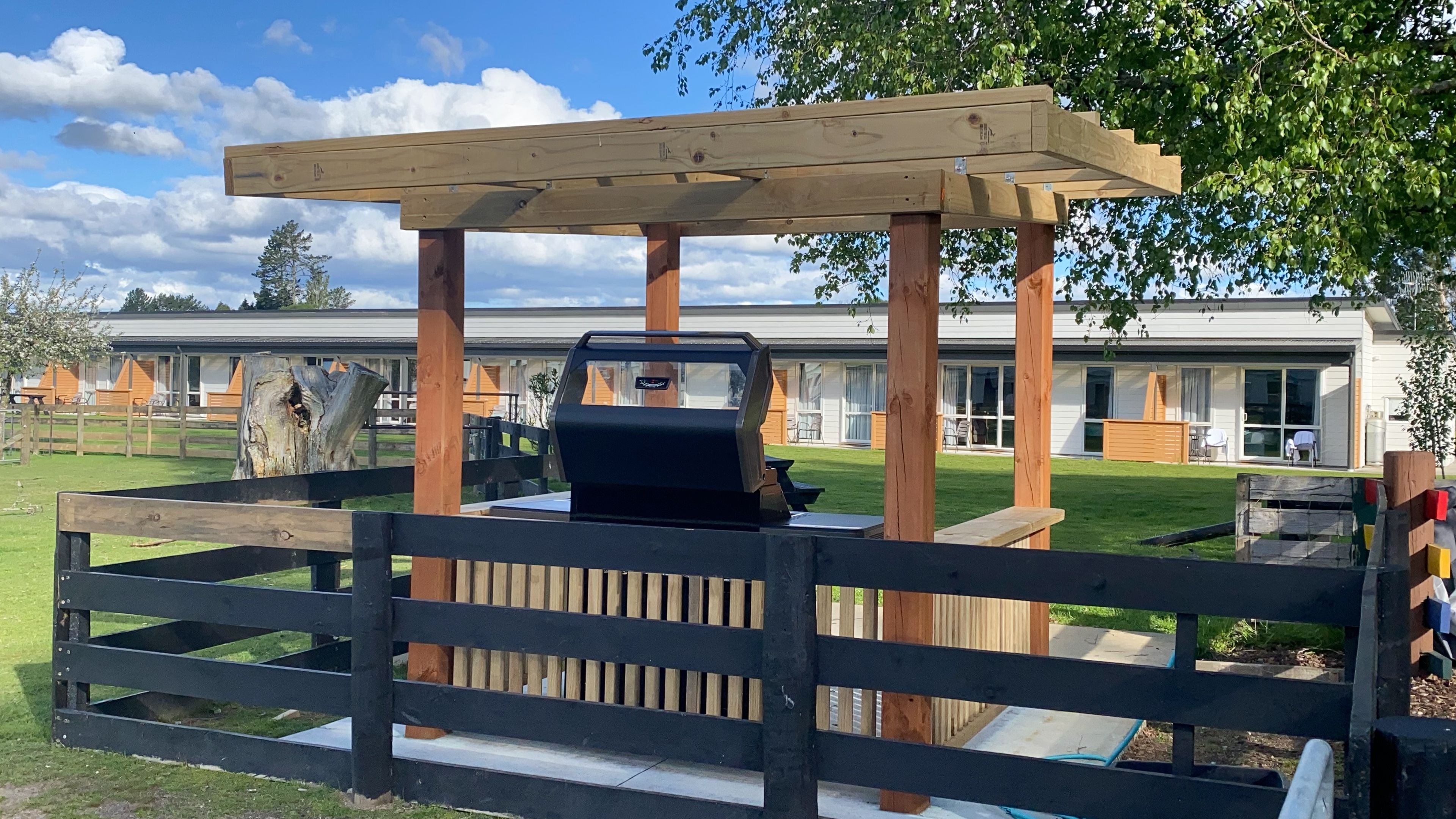 A wooden pergola with a barbecue grill underneath, surrounded by a wooden fence, with a building in the background.