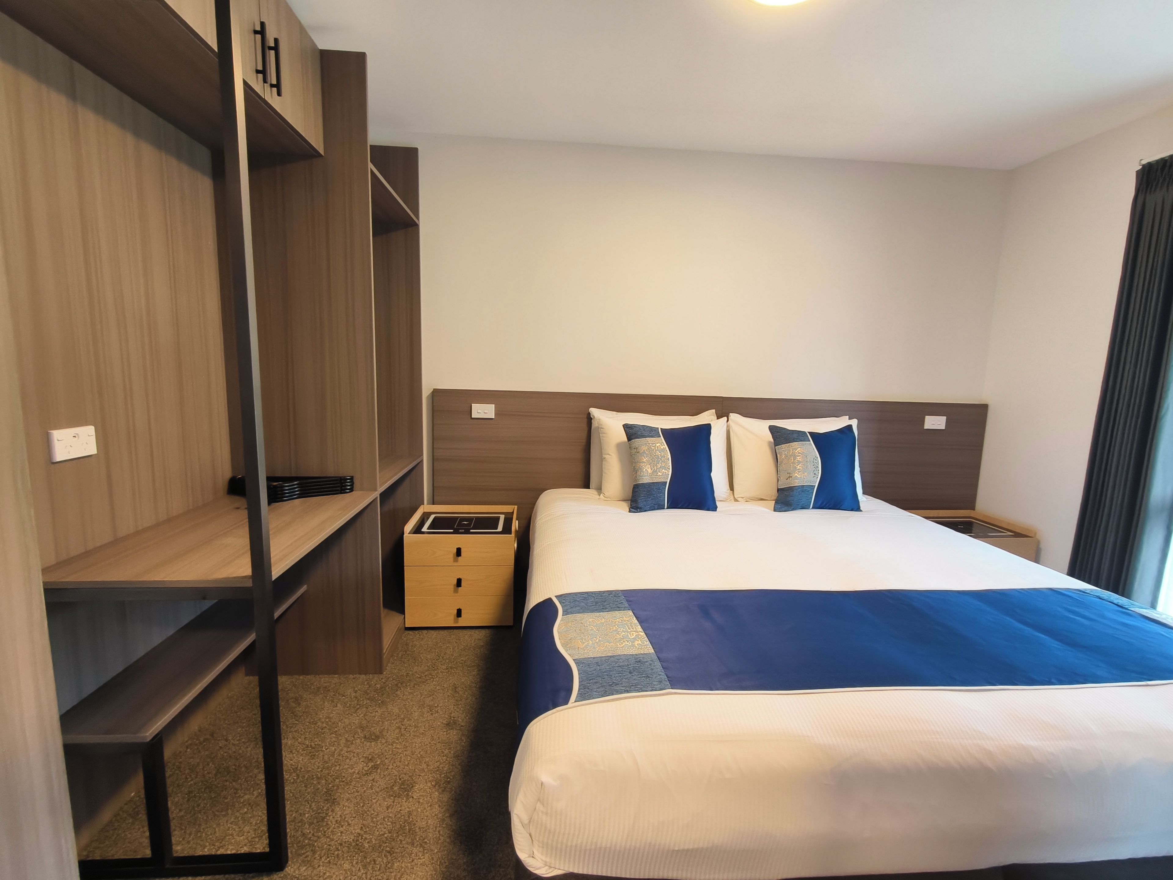 A hotel room with a large bed, a wooden wardrobe, a bedside table, and a small drawer unit.