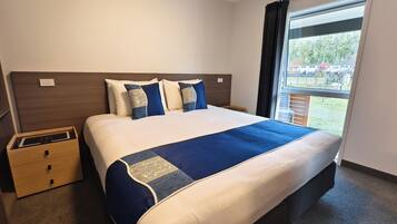 A hotel room with a large bed, a nightstand, a wooden drawer, and a window with a view of the outdoors.