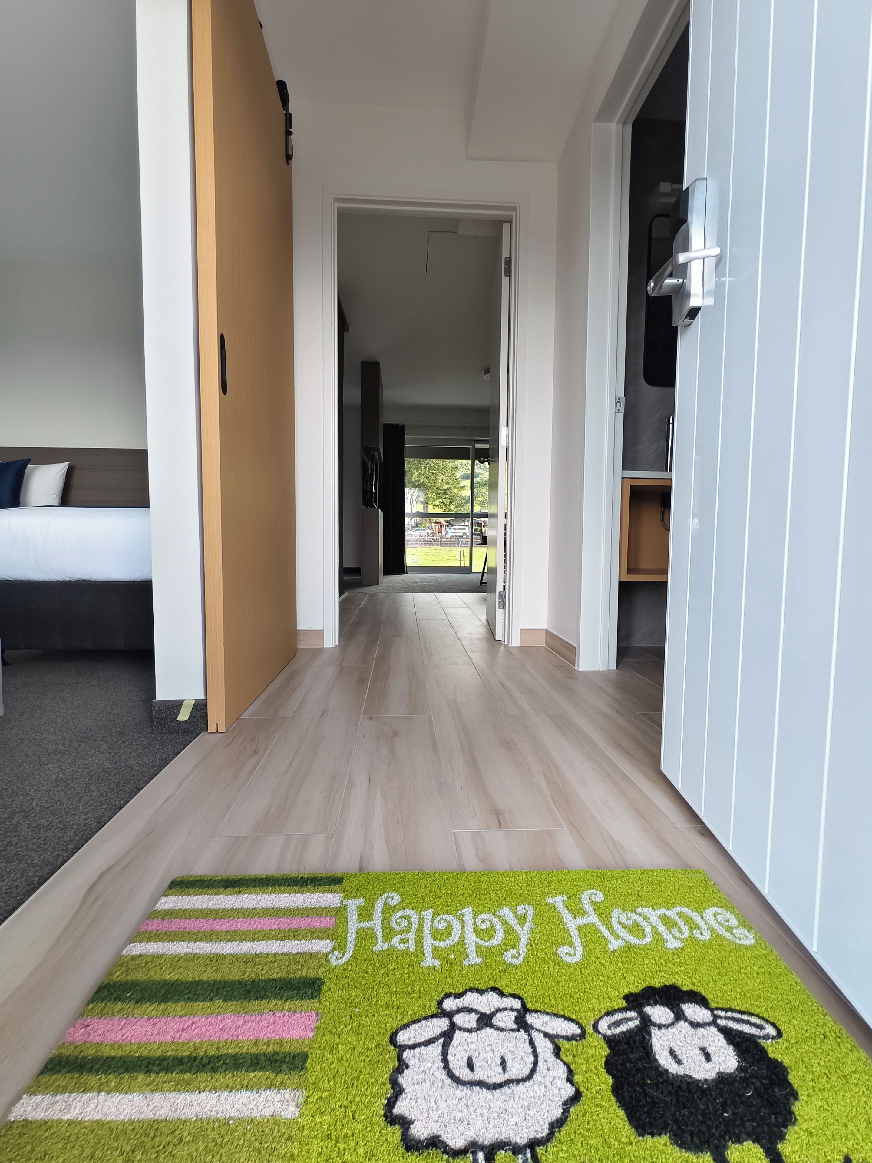 A hotel hallway with a carpet featuring a 'Happy Home' design, a bed with a dark headboard, and a door leading to a room with a view.