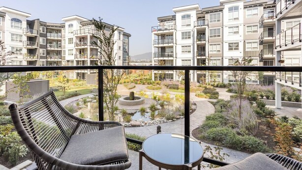Un balcón con mobiliario de mimbre, una mesa pequeña y vistas a un jardín ajardinado con un estanque y edificios al fondo.