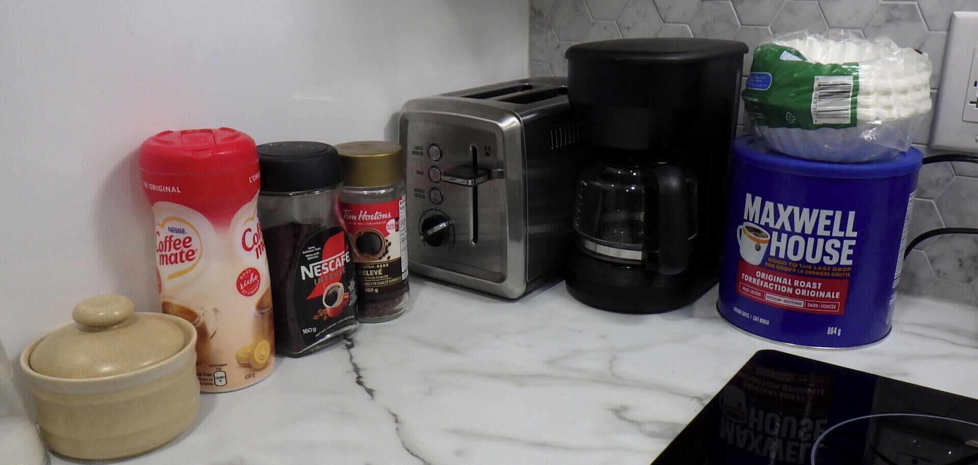 Eine Küchenarbeitsplatte mit Kaffeemaschine, Toaster, Kaffeemühle und einer Kaffeekanne Maxwell House, sowie ein Gläschen Kaffeeweißer.
