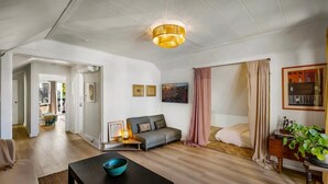 Un salón espacioso con un sofá, una mesa de centro y una araña. Se ve una puerta que conduce a otra habitación, y una planta a la derecha.