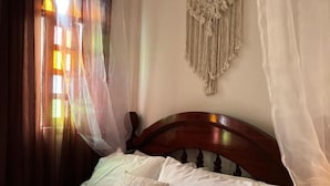 A bed with white linens and a wooden headboard, a macrame wall hanging, and sheer curtains allowing sunlight to filter through.