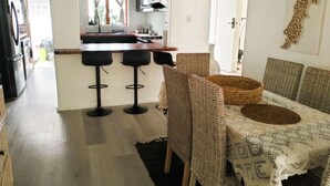 Een eetgedeelte met rieten stoelen en een tafel, een keuken met een centraal eiland en barstoelen, en een uitzicht door een boog naar een andere kamer.