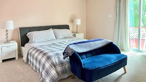 Un dormitorio con una cama, dos mesitas de noche con lámparas, una silla azul, una ventana con cortinas e iluminación empotrada en el techo.
