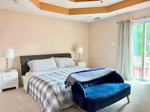 Een slaapkamer met een bed, twee nachtkastjes met lampen, een blauwe stoel, een raam met gordijnen en een plafond met inbouwverlichting.