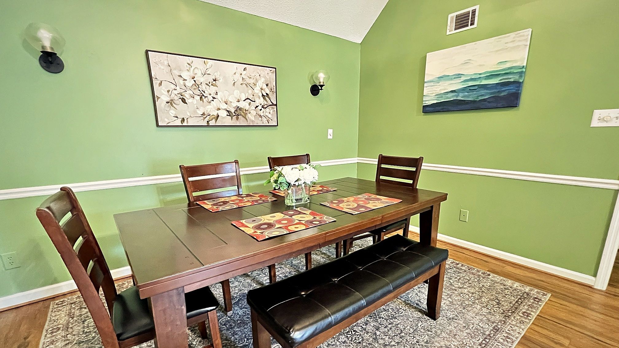 A dining room with a wooden table, four chairs, a bench, a vase with flowers, and two wall-mounted light fixtures.