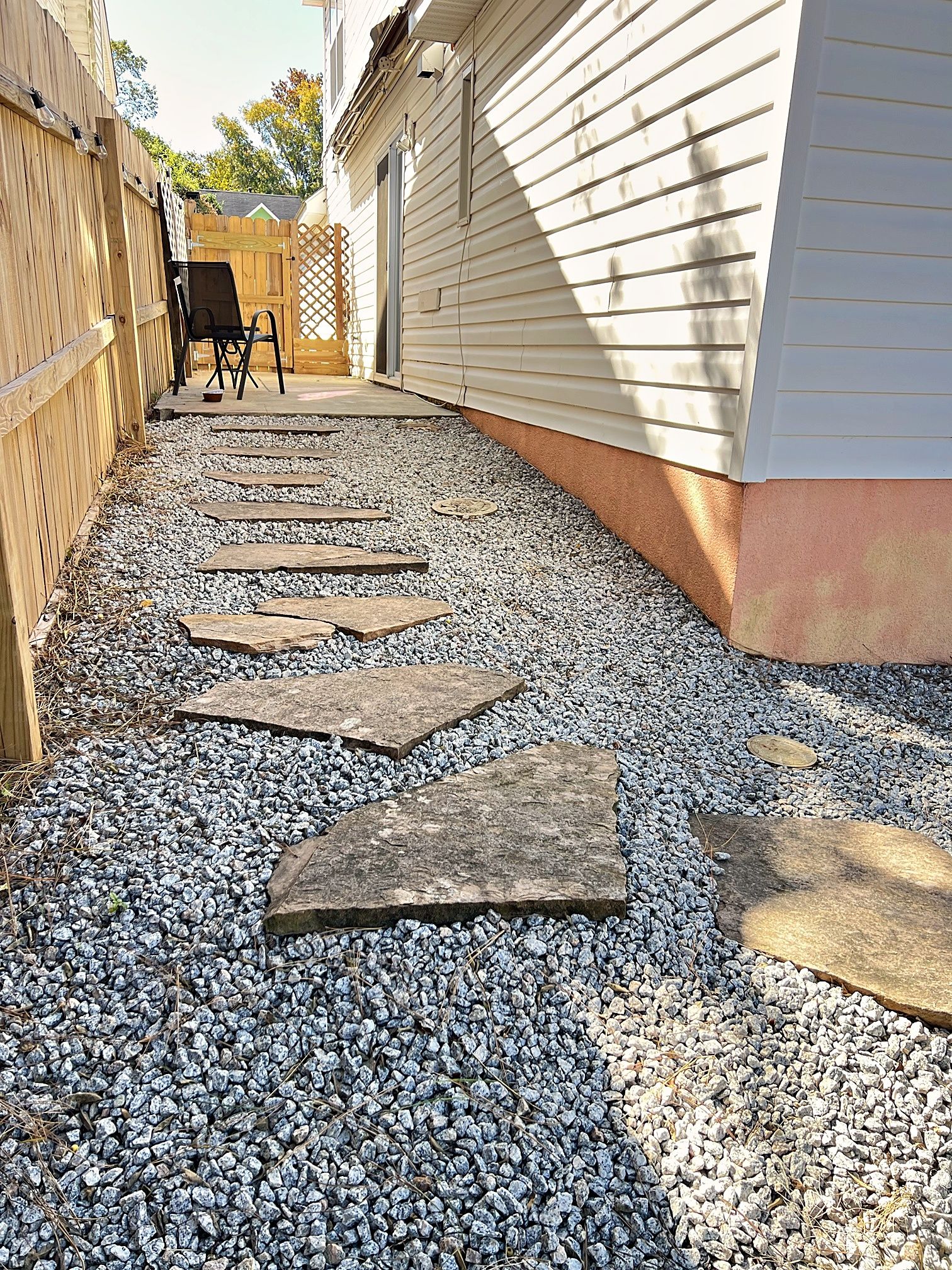 A gravel pathway leading to a wooden deck with a chair and table, surrounded by a wooden fence and a house with siding.