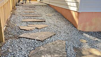 A gravel pathway leading to a wooden deck with a chair and table, surrounded by a wooden fence and a house with siding.