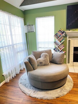 Un salon avec un canapé rond de couleur grise, un tapis blanc et moelleux, une cheminée et une bibliothèque remplie de livres.