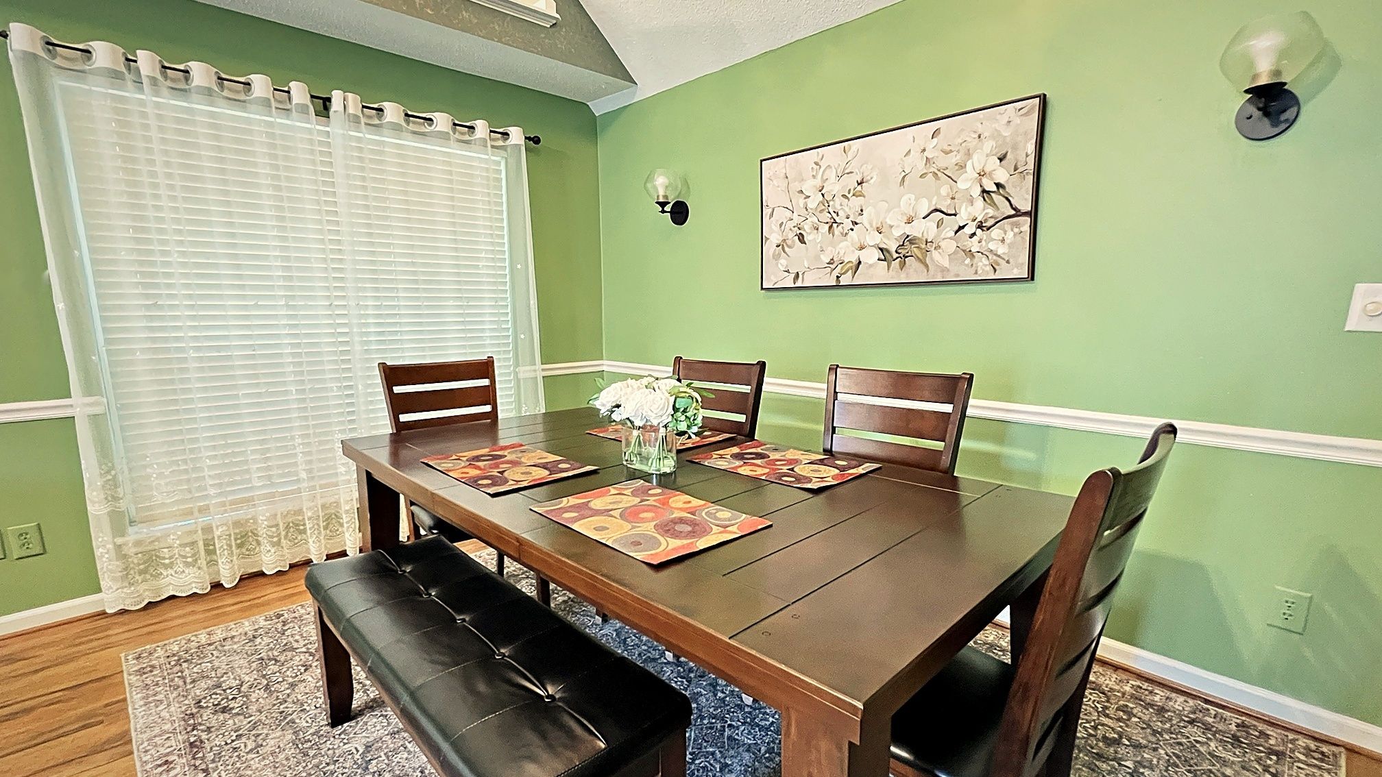 A dining room with a wooden table, four chairs, a bench, a vase with flowers, and a wall-mounted light fixture.