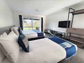 Habitación de hotel con dos camas, un escritorio, una silla, un televisor y una ventana con vista a un edificio y una valla.