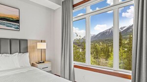 Een hotelkamer met een bed, nachtkastje met een lamp, en een groot raam met uitzicht op bergen.