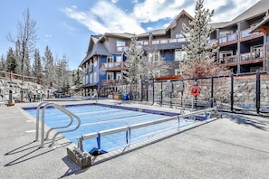 A pool area with a clear sky, a stone wall, and a building with multiple balconies. - Scenic 3BR Retreat with MTN Views, Hot Tub and Pool (Canmore)