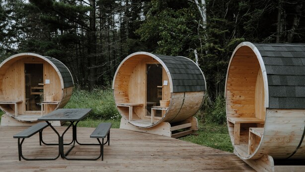 Three wooden barrel-style cabins with attached benches and a picnic table in a forest setting.