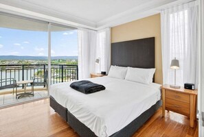 A hotel room with a large bed, a wooden nightstand, a desk with a chair, and a balcony with a view of the city. - Towers of Chevron - 3122 (2BR/1BA) (Gold Coast)