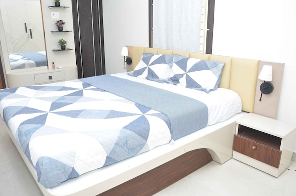 A neatly made bed with a geometric patterned bedspread, a bedside table with a lamp, and a small shelf with decorative items.