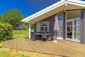 A covered outdoor deck with a table and chairs, large windows, and a view of a house in the background. - Holiday home with a spa and sauna by the water (Rudkøbing)
