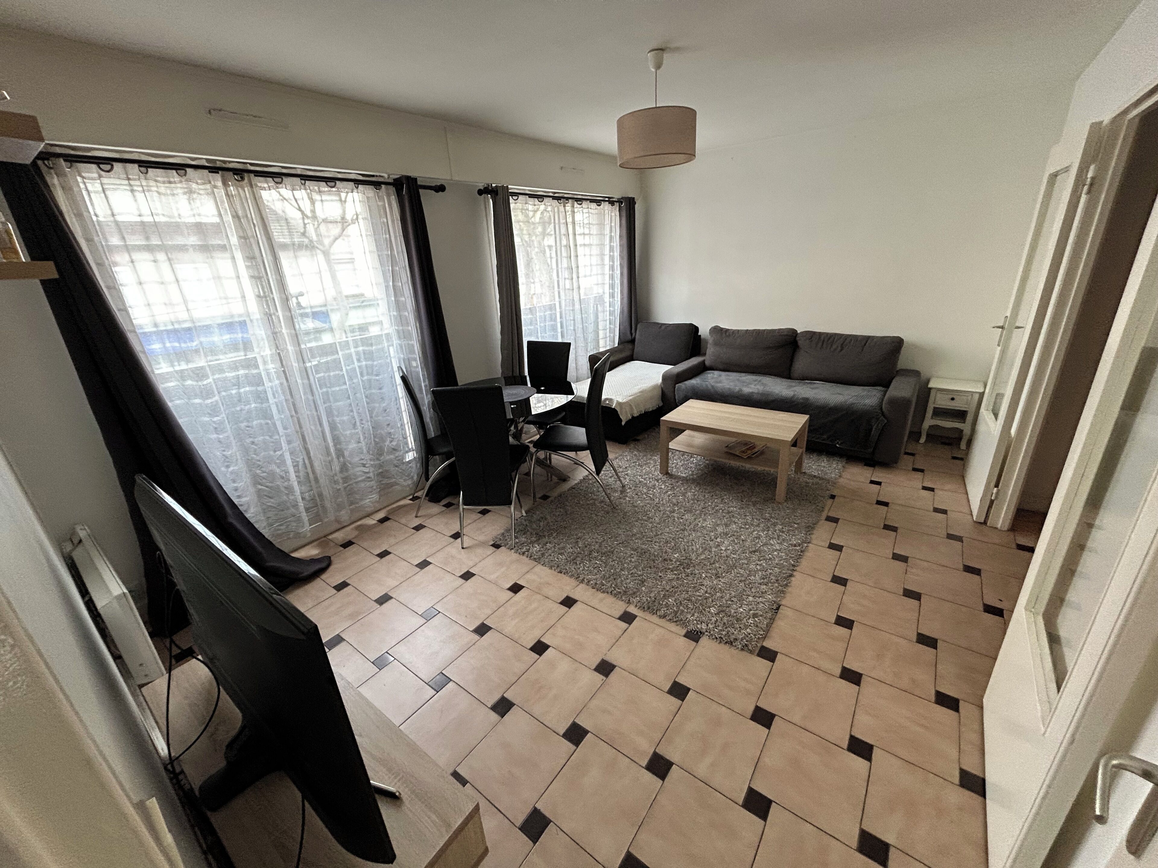 Un salon avec un canapé, une table basse et un téléviseur. Il y a une fenêtre avec des rideaux, un sol carrelé et une porte.