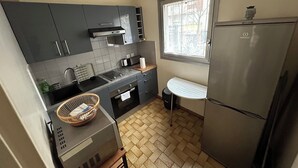 A compact kitchen with grey cabinets, a stainless steel refrigerator, a small white table, and a window with a view of the outside. - Lovely Family Stay - Two  Bedroom - Right Edge of Paris (Clichy)