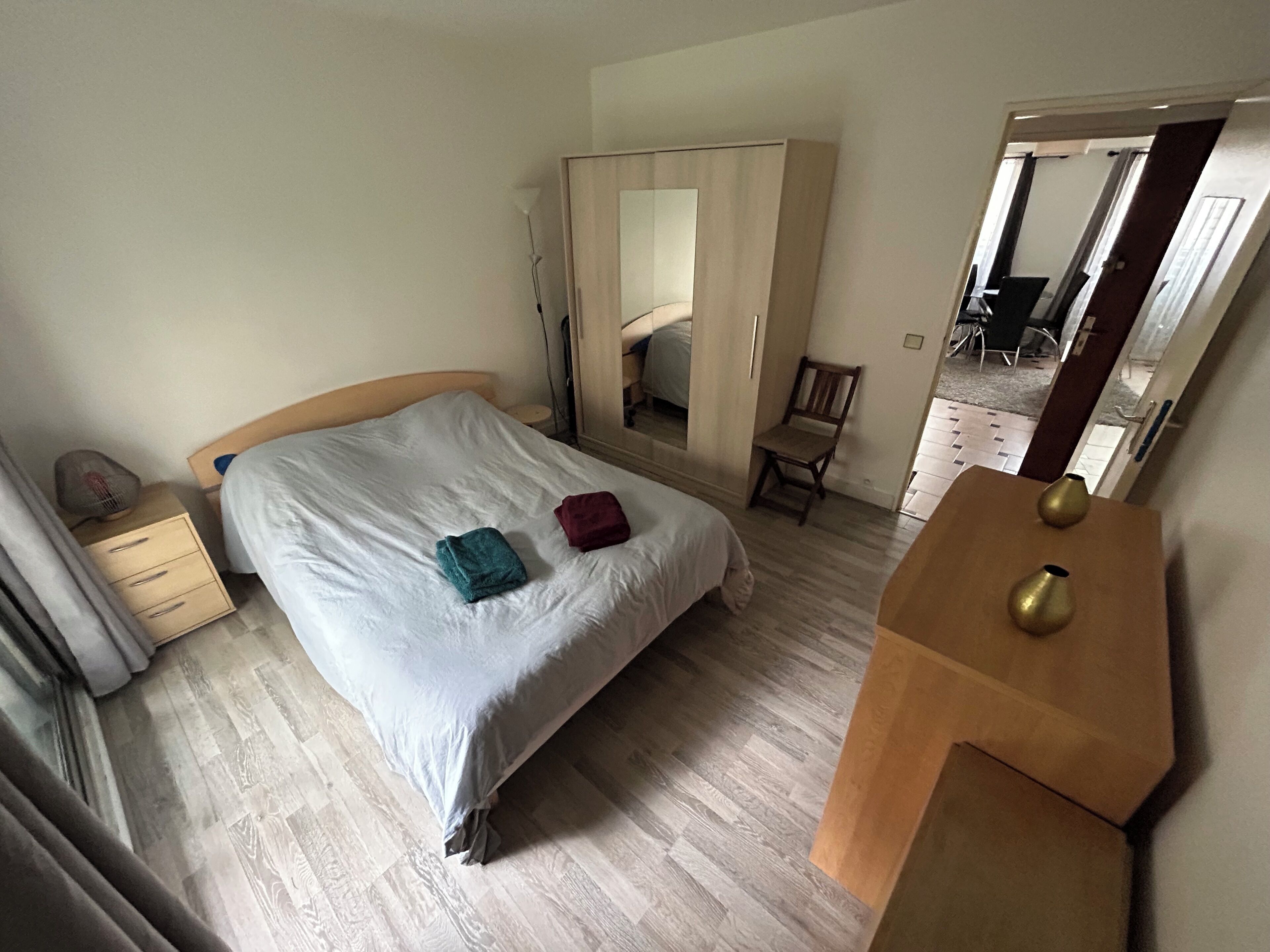 A bedroom with a bed, a wooden nightstand, a chair, a wardrobe, and a mirror.