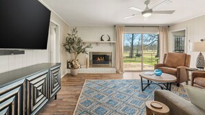 A living room with a fireplace, a flat-screen TV, a ceiling fan, and a view of the outdoors.