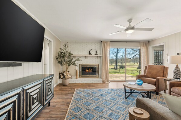 A living room with a fireplace, a flat-screen TV, a ceiling fan, and a view of the outdoors.