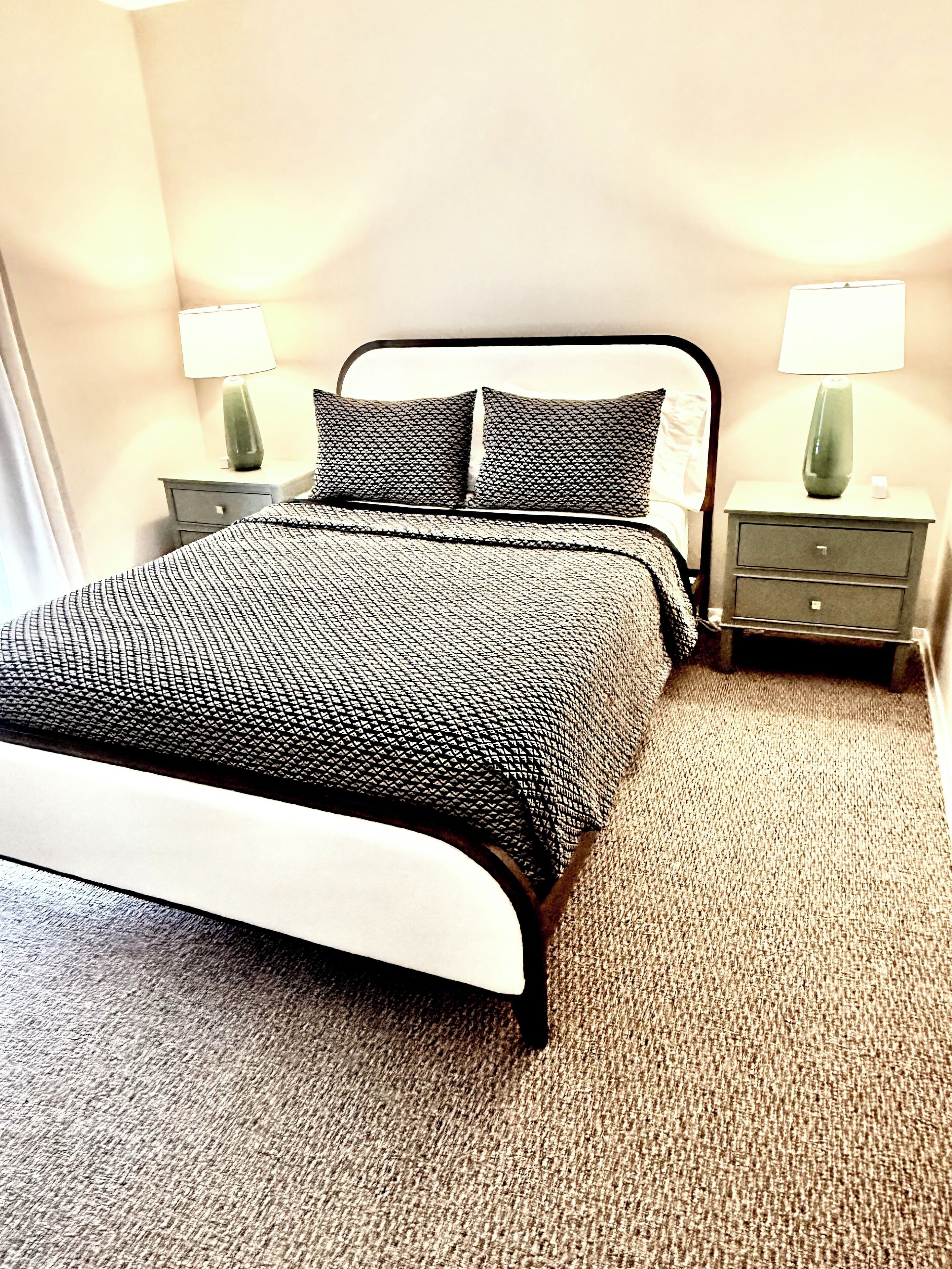 Un lit bien fait, recouvert d’une couvre-lit à motifs noir et blanc, deux tables de chevet avec des lampes et un sol recouvert d’une moquette.