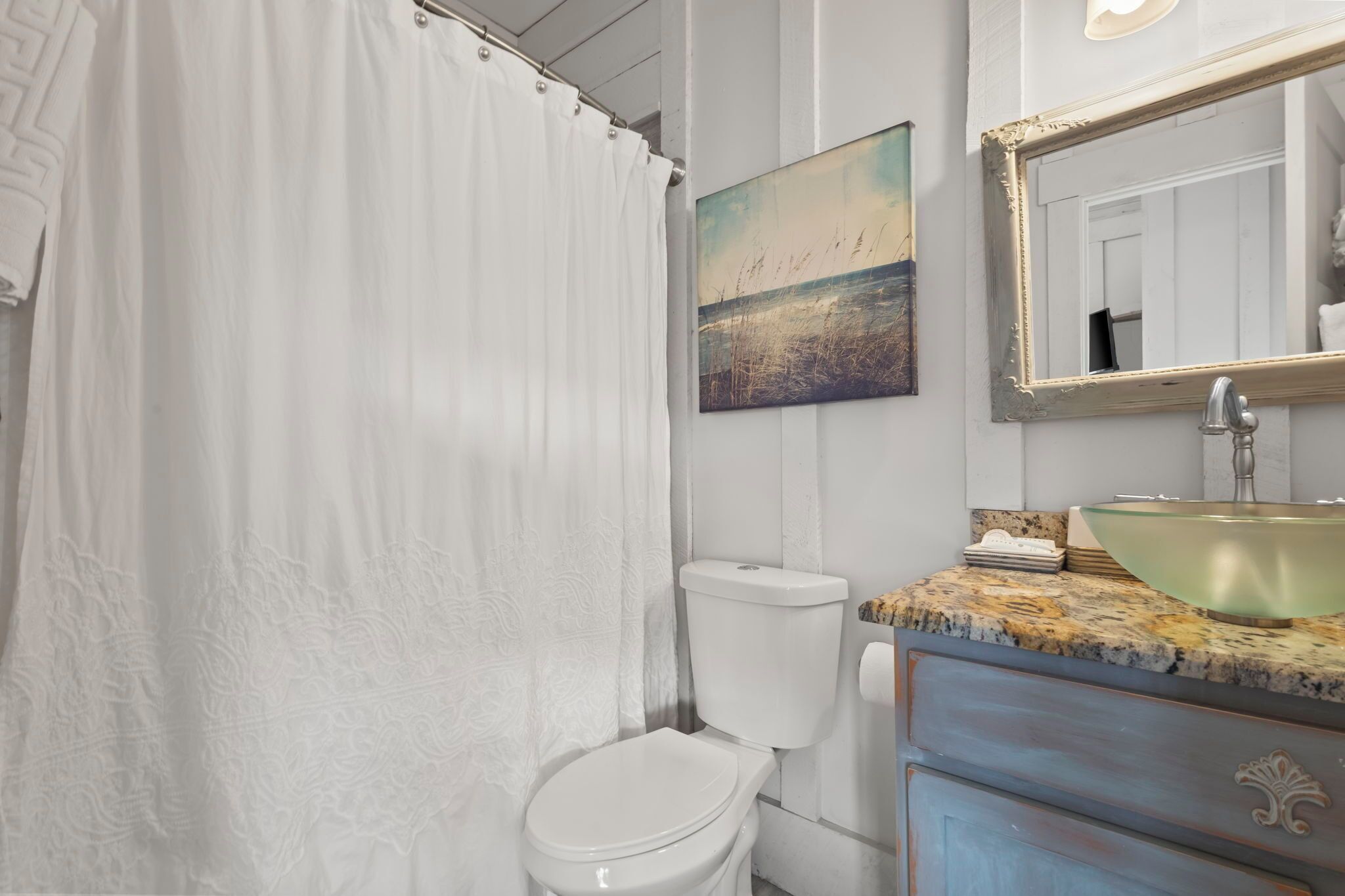 Un baño con inodoro blanco, lavamanos de vidrio y un cuadro de una playa.