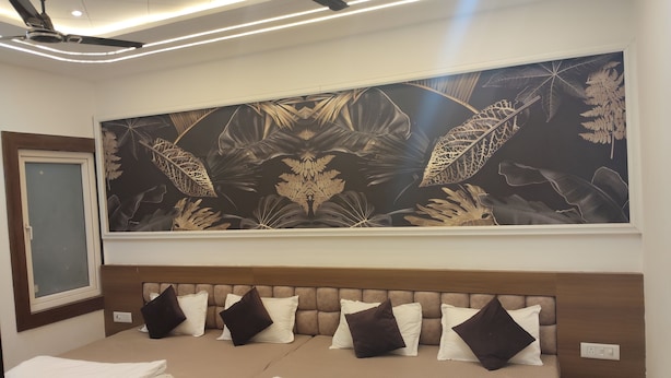 A hotel room with a bed, a large wall mural of tropical leaves, ceiling fans, and a window with a view. - Shri Ramya Dham (Mathura)