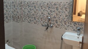 A bathroom with a toilet, green bucket, small stool, shower, and sink. - Shri Ramya Dham (Mathura)