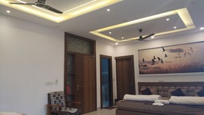 A modern living room with a sofa, a chair, and a bed. There are ceiling fans and recessed lighting. - Shri Ramya Dham (Mathura)