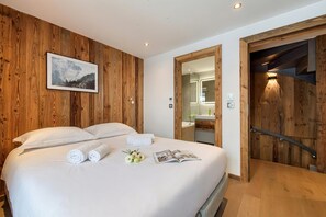 Ein ordentlich bezogenes Bett mit aufgerollten Handtüchern, ein gerahmtes Bild an der Wand und eine Holztür zu einem Badezimmer.