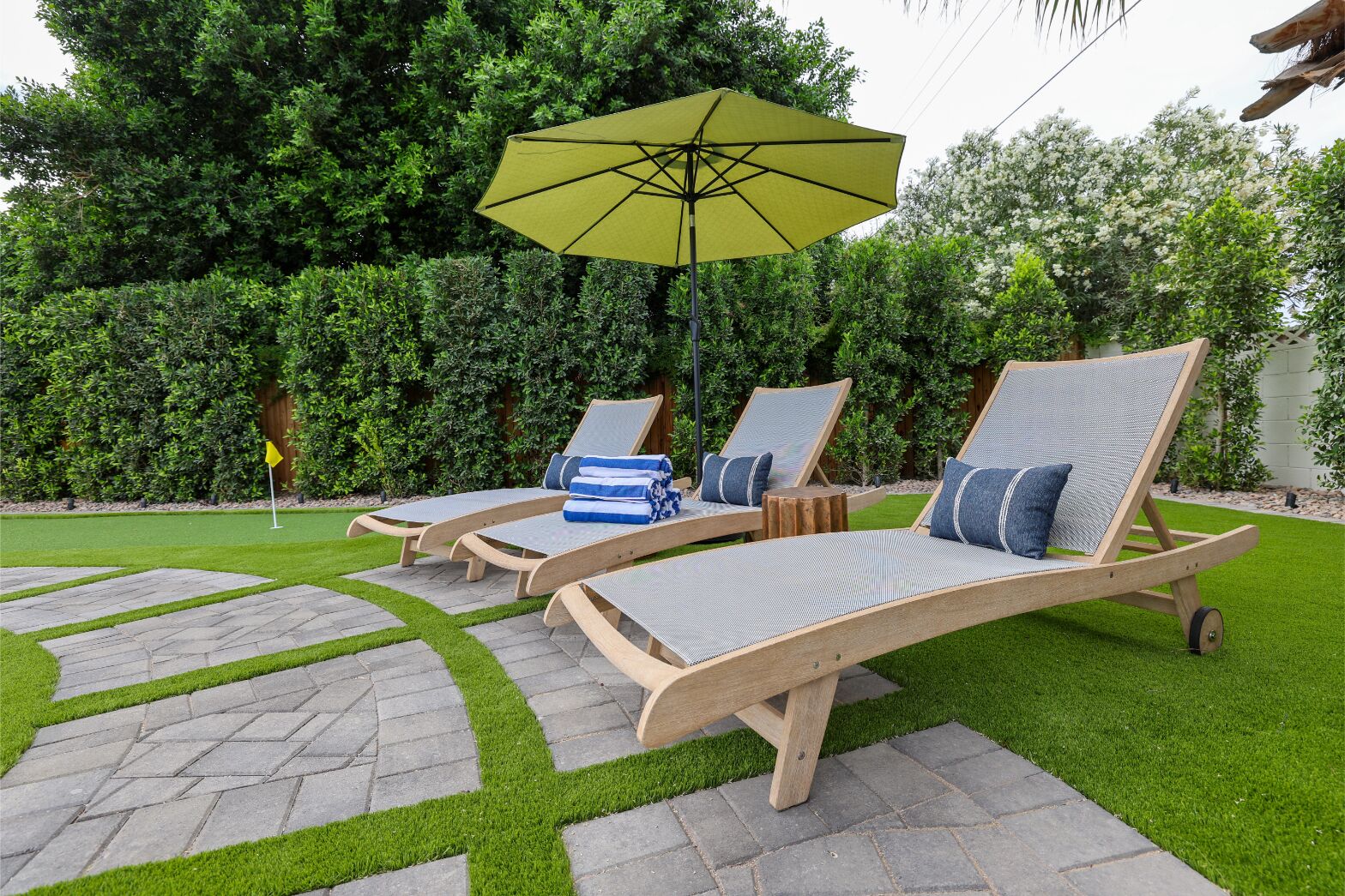 Three wooden lounge chairs with cushions, a yellow umbrella, and a flag on a well-maintained lawn with stone pathways.