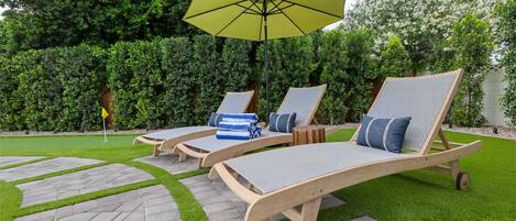 Three wooden lounge chairs with cushions, a yellow umbrella, and a flag on a well-maintained lawn with stone pathways.