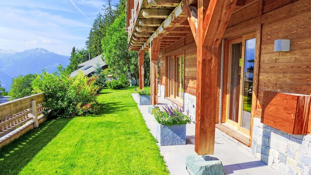 A wooden cabin with a stone foundation, a well-maintained lawn, and a view of mountains in the background.