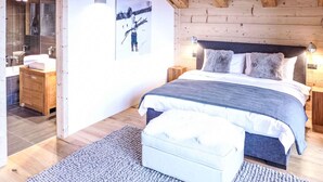 A bedroom with a wooden ceiling, a bed with a grey and white duvet, two bedside tables with lamps, a wooden nightstand, a grey rug, and a framed picture on the wall.