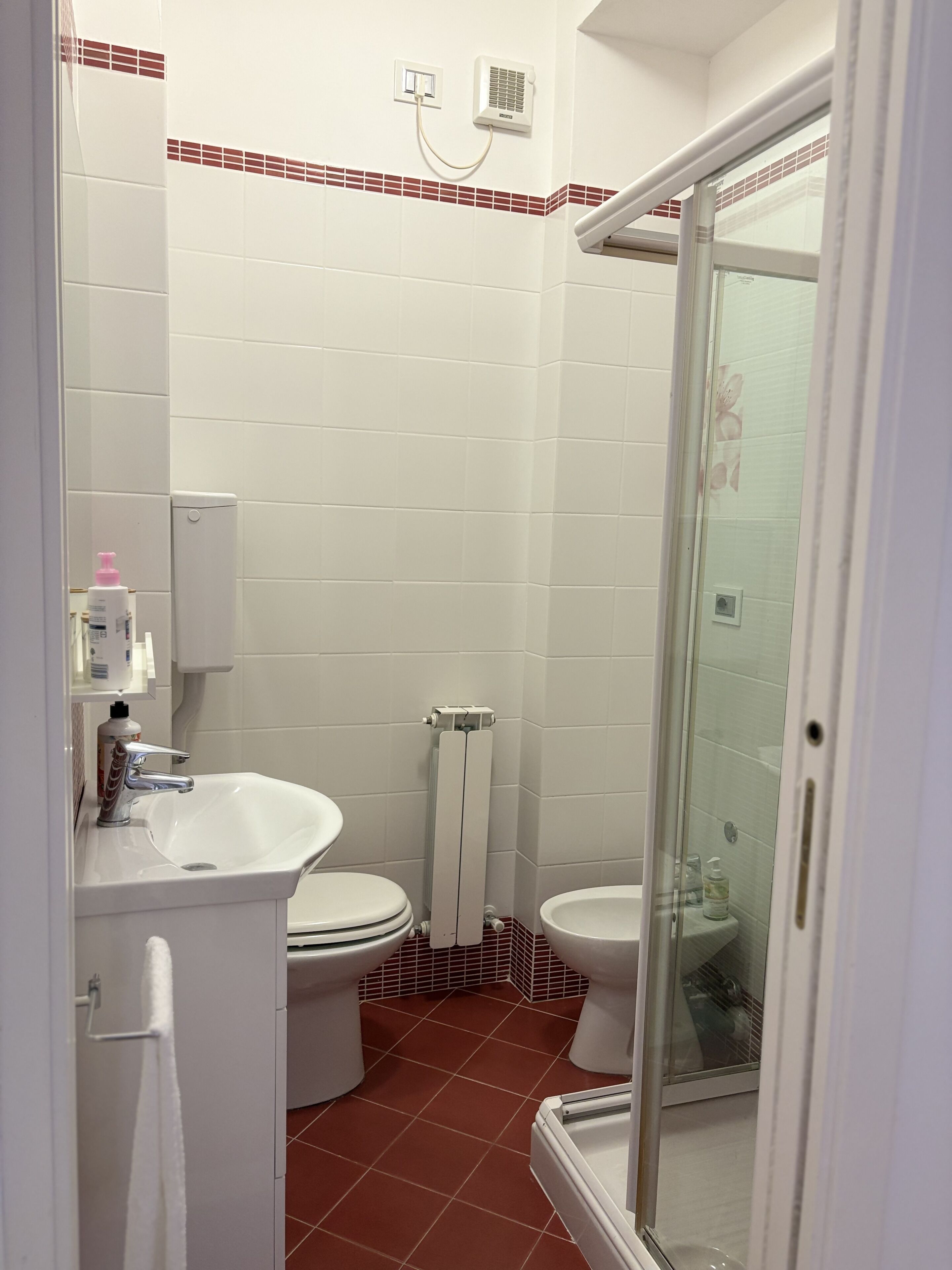 A bathroom with white tiles, a glass shower enclosure, a white sink, and two toilets.