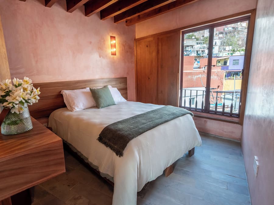 Un dormitorio con cabecera de madera, un ventanal con balcón y una mesita de noche con un jarrón de flores.