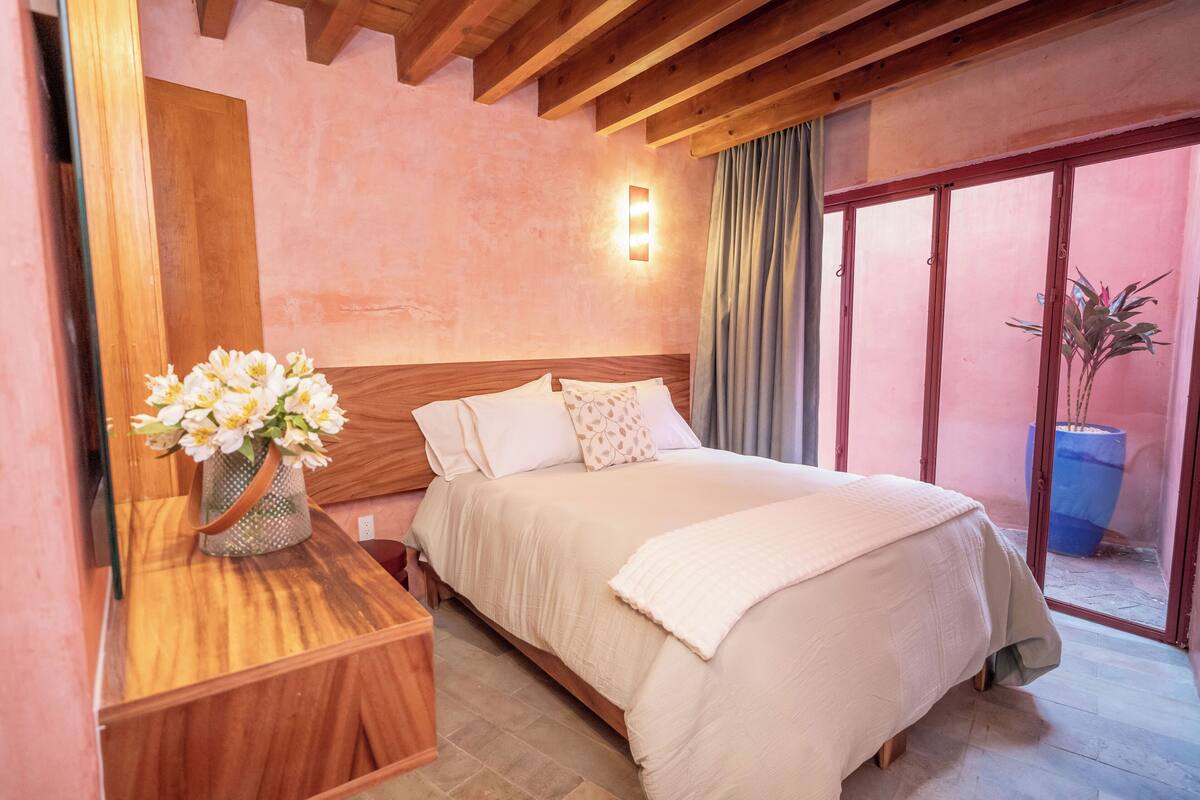 Ein Schlafzimmer mit einem hölzernen Kopfende, einem Nachttisch mit Blumen in einer Vase, einem Bett mit weißer Bettwäsche und einer Glastür, die zu einem Außenbereich mit einem Blumentopf führt.