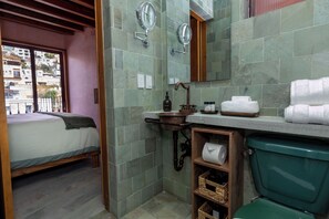 A bathroom with a green toilet, a wooden shelf with toiletries, a sink with a mirror, and a bed in the adjacent room. - Hotel Casa de las Abuelas (Guanajuato)