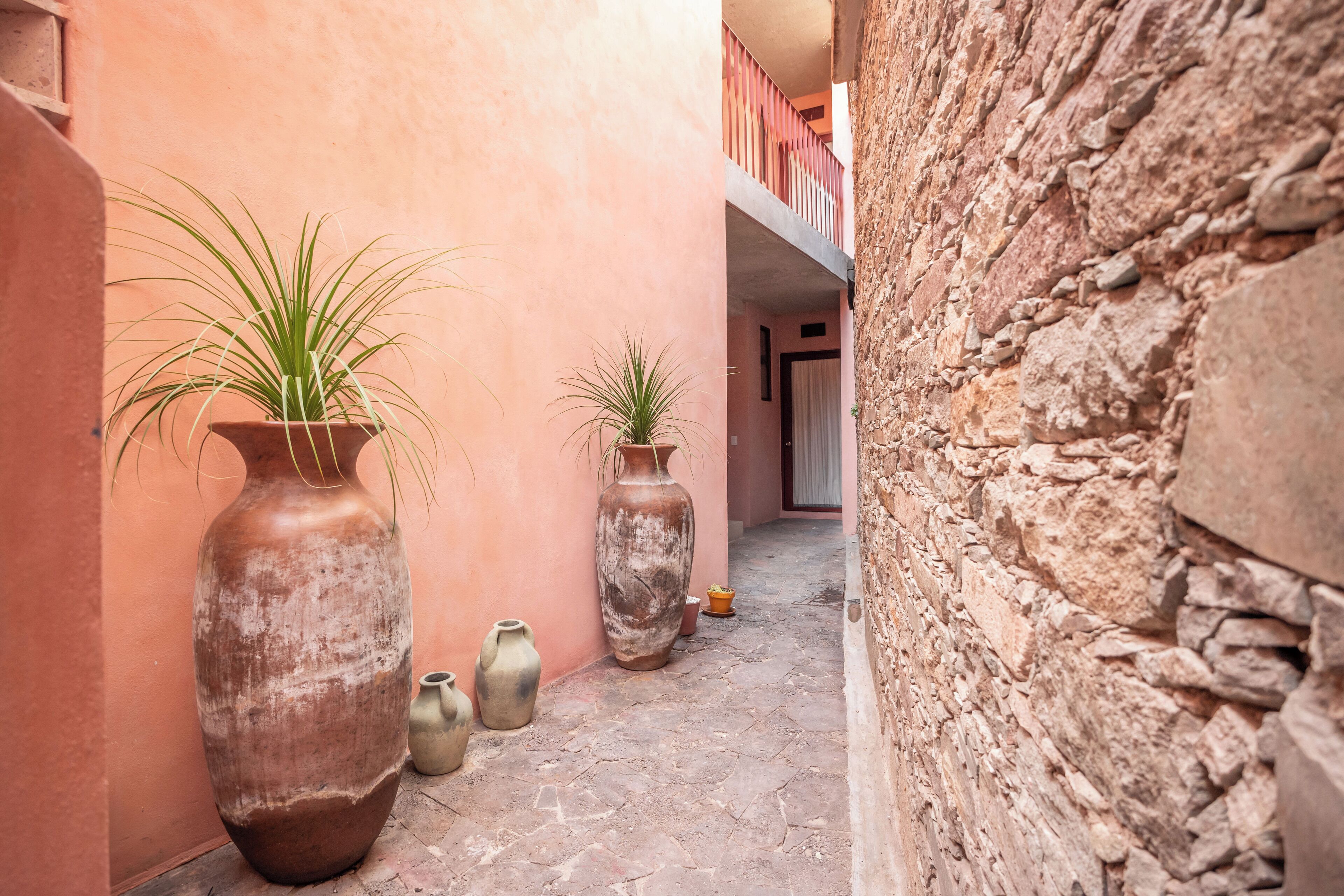 A narrow alley with large vases and potted plants, stone walls, and a balcony.