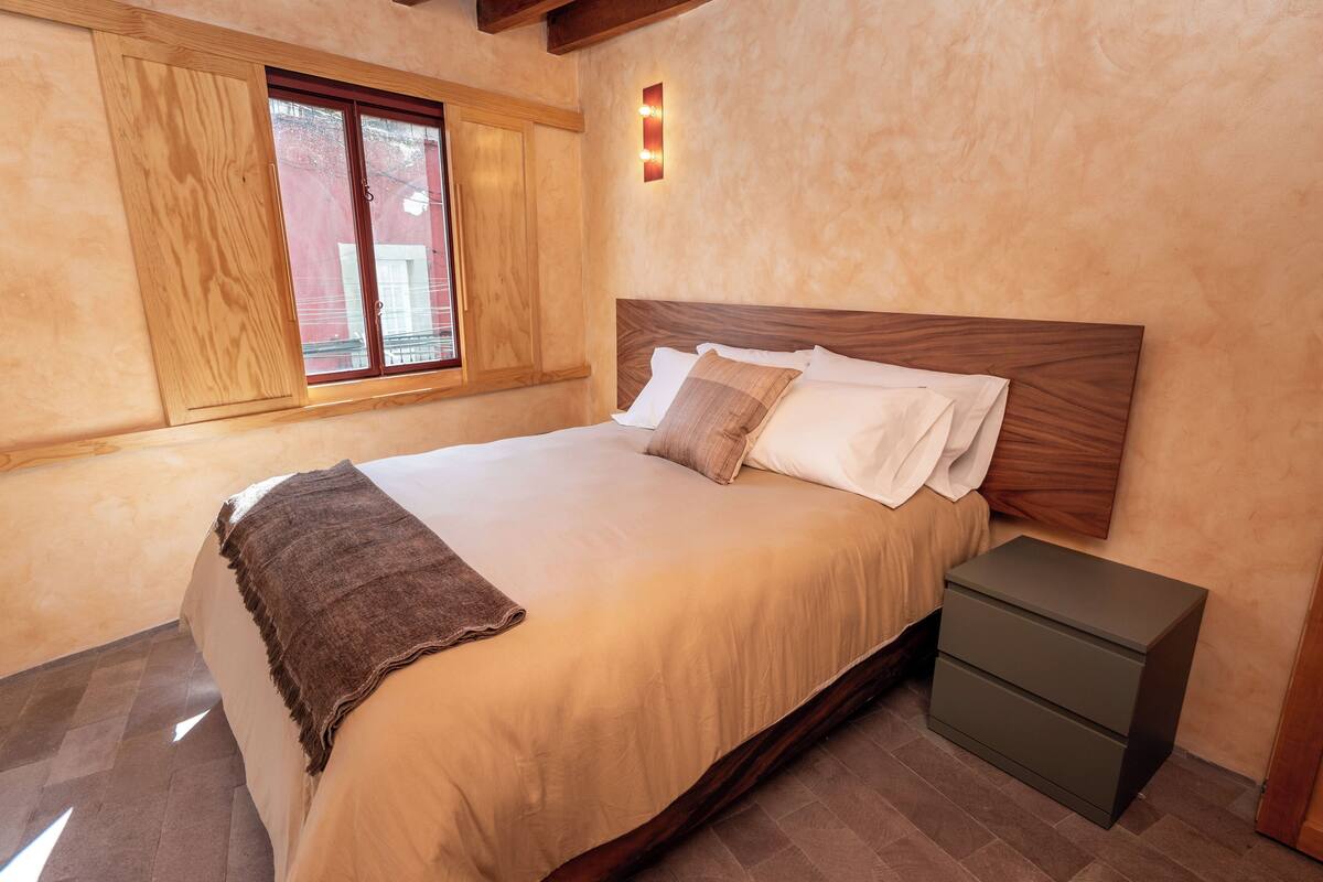 Ein ordentlich bezogenes Bett mit Holz Kopfteil, einem Nachttisch und einem Fenster mit Holzjalousien.