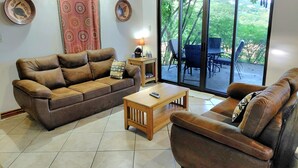 Een woonkamer met een bruine leren bank, een houten salontafel en uitzicht op een terras met tuinmeubilair.