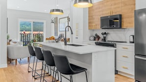 A modern kitchen with a central island, bar stools, a built-in oven, and a large window offering a view of the outdoors. - 4BR Getaway with MTN View, Balcony Near DT & Banff! (Canmore)