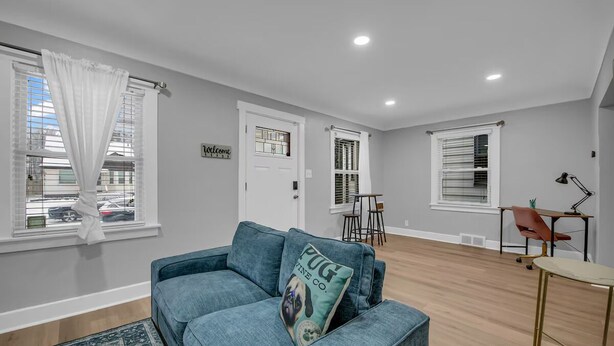 A modern living room with a blue sofa, a small coffee table, and a rug. There are two windows with blinds, a door, and a desk with a chair. - Cozy Home Near Ferndale, Royal Oak & Detroit! (Hazel Park)