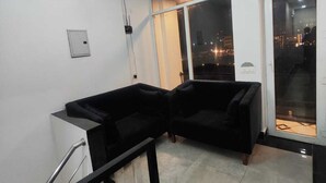 A modern living room with a black sofa set, a staircase, and a large window offering a city view. - BLACKSTORM RESIDENCY HOTEL (Greater Noida)