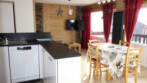 A kitchen with a black countertop, white cabinets, and a built-in dishwasher. There is a dining area with a table covered by a tablecloth with a pattern, and wooden chairs. A television is mounted on the wall above the kitchen. - Apartment in France Near Ski Lifts (Huez)