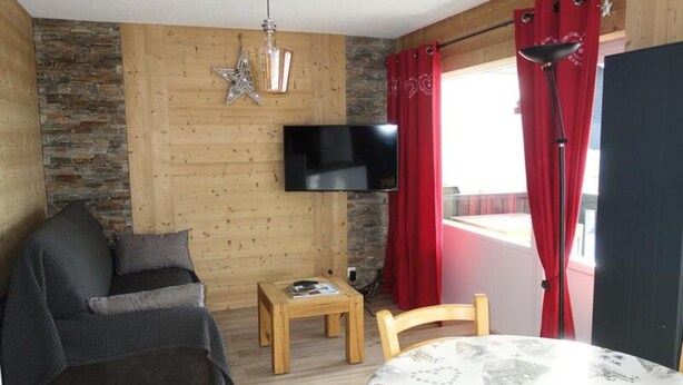 A cozy living room with a stone wall, a wooden ceiling, a flat-screen TV, a small wooden table, a grey sofa, and a red curtain. - Apartment in France Near Ski Lifts (Huez)